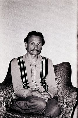 Portrait of a man in a relaxed pose, seated on a plush armchair with vertical striped shirt and buttoned collar. Mid-century ...