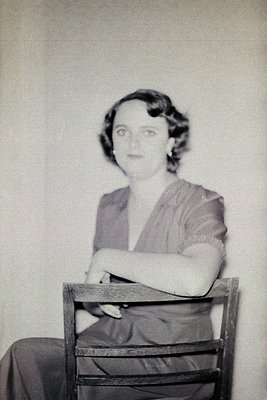 Portrait of a woman in a 1930s–1940s dress with a fitted bodice and short sleeves, seated on a wooden chair against a plain w...