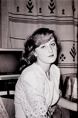 Portrait of a woman in a 1950s–1960s indoor setting, wearing a lace-up blouse with a high-neck collar. Geometric-patterned wa...