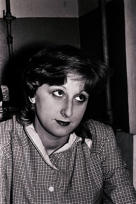 Portrait of a woman in a checkered blouse, likely 1960s–1970s. Indoor setting with industrial pipes and lighting fixtures. Ca...