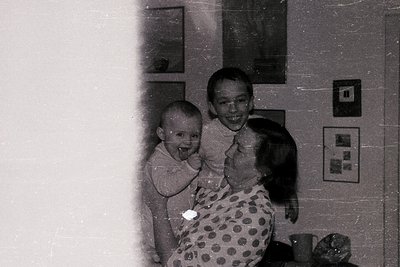 Vintage black-and-white photo of three generations indoors, likely mid-20th century. A toddler in a short-sleeved shirt, an a...