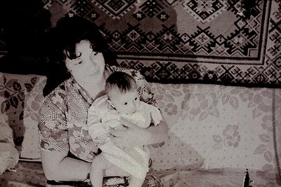 Vintage black-and-white photo of a woman cradling an infant on a patterned sofa, likely mid-20th century. Geometric and flora...