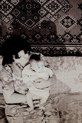 A tender black-and-white moment of a woman cradling an infant on a floral-patterned sofa, adorned with geometric wallpaper. L...