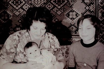 Vintage black-and-white photo of a woman cradling a baby, with a young child seated beside her. Patterned wallpaper features ...