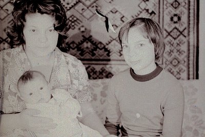 Family portrait from the 1960s–1970s featuring a woman holding a baby, with a young child beside her. Patterned wallpaper wit...