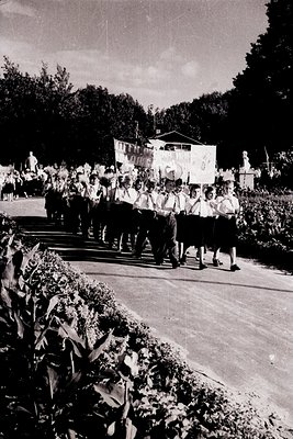 Group of uniformed youth marching in formation through a landscaped park, likely a 1950s–1960s Soviet-era event. Uniforms fea...