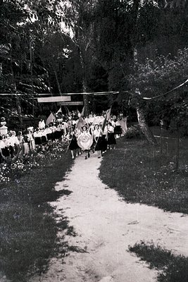 Vintage parade procession featuring uniformed groups marching on a dirt path through a wooded area, likely 1950s–1960s. Flags...