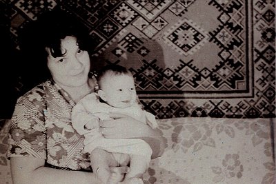 Vintage black-and-white portrait of an adult holding a baby indoors, likely mid-20th century. Geometric patterned wallpaper a...