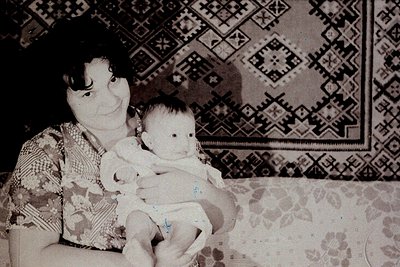 Vintage black-and-white portrait of a woman cradling an infant, set against a patterned wall and floral upholstery. Geometric...