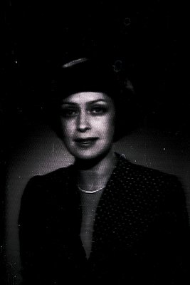 Vintage black-and-white portrait of a woman in mid-20th century fashion, likely 1940s–1950s. She wears a fitted jacket with a...