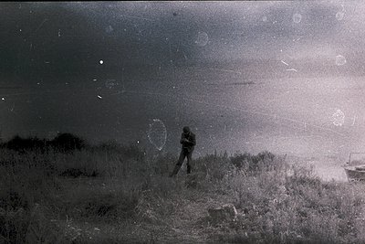 Vintage black-and-white photo of lone figure in overcoat standing on windswept grassy dune, facing ocean horizon. Boat partia...