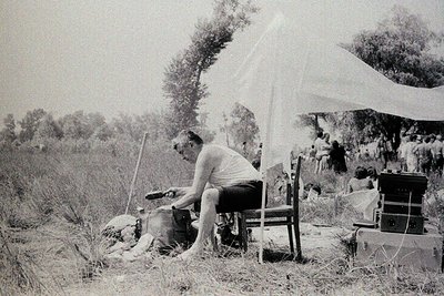Black-and-white snapshot of a man sharpening a tool at an outdoor campsite, seated on a wooden bench. Surrounding him: a dog ...