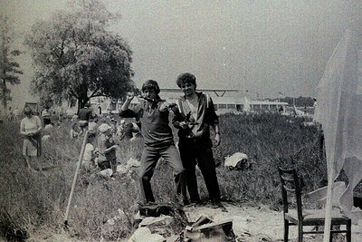 Black-and-white photo captures two young men in mid-1970s Eastern Europe, posing on a makeshift wooden platform amid construc...