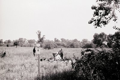 Two men in rural field near "Private" sign, mid-20th century. One kneels beside a prone figure, possibly a casualty. Dense fo...