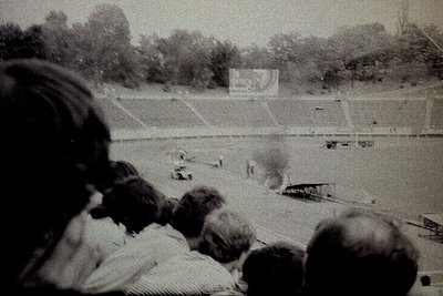 Black-and-white stadium shot showing a mid-20th century track event, likely a decathlon or pentathlon. Crowd in foreground, b...