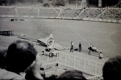 Olympic torch relay procession with flame in stadium, mid-20th century. Crowd watches from stands as torchbearers carry flame...