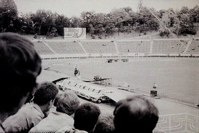 Black-and-white shot of a vintage airshow at a stadium, featuring vintage aircraft on display. Crowd in foreground watches pi...