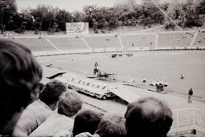 Aerial view of a mid-20th-century stadium event, likely a 1960s/1970s motorsport race. Spectators in tiered seating watch a v...