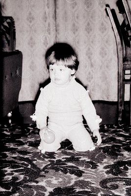 Toddler in vintage-style onesie kneeling on patterned carpet, holding a small rattle. Mid-20th century domestic interior with...