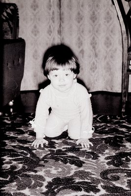 Vintage black-and-white photo of a toddler crawling on patterned carpet, mid-20th century. Indoor setting with floral wallpap...