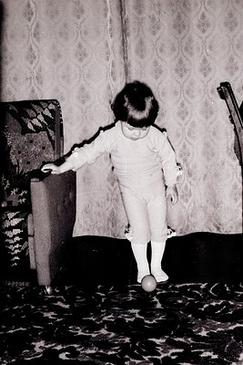 A toddler in a white onesie and white socks balances on a single ball in an indoor setting, likely mid-1970s. Patterned wallp...