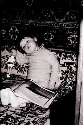 Vintage black-and-white portrait of a young child seated on an ornate, patterned sofa, engrossed in a book. The child wears a...