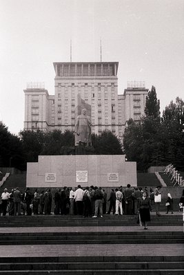 Monumental statue of a figure in military attire atop a stone pedestal, flanked by plaques, set in an urban plaza. Multi-stor...