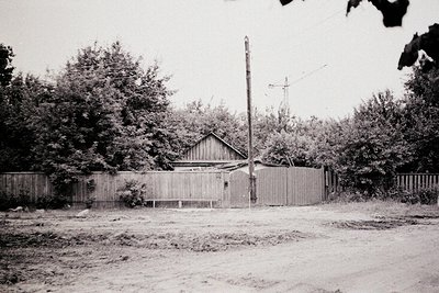 Mid-20th century rural home with wooden fence and utility pole. Single-story house with gabled roof, surrounded by dense foli...