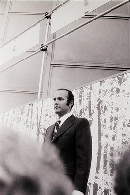 Man in a dark suit and patterned tie speaking indoors, likely at a conference or formal event. Industrial lighting and scaffo...