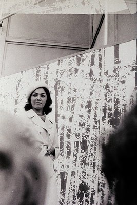 Black-and-white portrait of a woman in a tailored blazer and headscarf, standing in front of a patterned wall with geometric ...