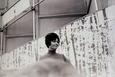 Black-and-white portrait of a woman in a turtleneck sweater, standing in front of a patterned wall with geometric designs. Th...