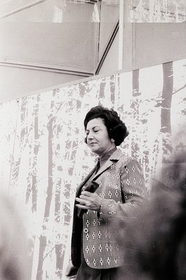 Mid-century woman in a patterned blouse and structured jacket poses indoors, likely a public or institutional setting. The fl...