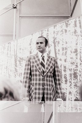 Portrait of a man in a **plaid blazer** and **white dress shirt**, standing in a **reflective glass-walled space** (likely an...