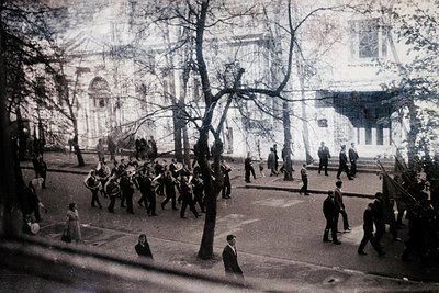Black-and-white street scene featuring a military parade with uniformed soldiers marching in formation, led by a band. Civili...