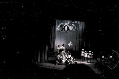 Vintage black-and-white stage performance featuring a dramatic curtain backdrop with "50th" anniversary emblem. Group of perf...