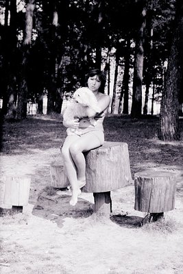 Black-and-white portrait of a woman seated on a tree stump in a forested area, cradling a small dog. Her pose suggests a cand...