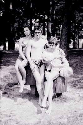 Three individuals pose casually on a wooden stump in a wooded area, mid-20th century. The man wears glasses and a patterned s...