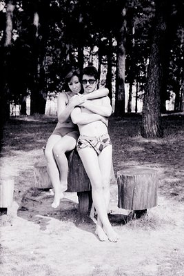 Two individuals pose playfully outdoors in a mid-20th-century beach setting, likely the 1960s. One wraps arms around the othe...