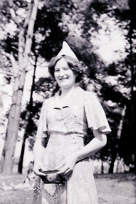 Vintage black-and-white portrait of a woman in early 20th-century outdoor attire, holding a large woven basket. She wears a w...
