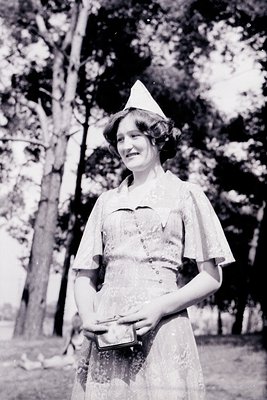 Vintage black-and-white portrait of a woman in a nurse’s uniform with a white cap, holding a small metal tray. The uniform fe...