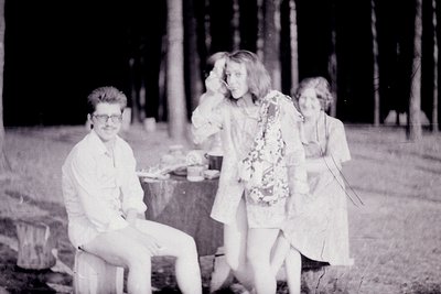 Vintage black-and-white photo of three individuals in outdoor setting, likely mid-20th century. Man sits on bench in short-sl...