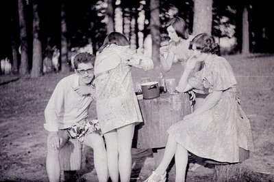 Vintage black-and-white photo of three young people in a forested area, mid-20th century. A man in glasses and a short-sleeve...