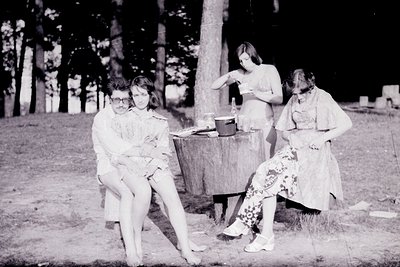 Four women in 1970s outdoor setting, likely a rural or suburban backyard. Two seated on grass, one holding a child, another s...