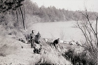 Black-and-white image of two men by a serene lake, likely mid-20th century. One man stands holding a fishing rod; the other s...