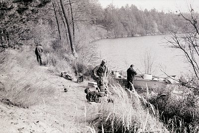 Black-and-white scene of three men near a frozen lake, mid-20th century. Two men load supplies onto a small boat on the shore...