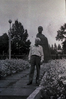 A man in a light button-down shirt and trousers stands beside a large, imposing statue of another figure in a park setting. T...