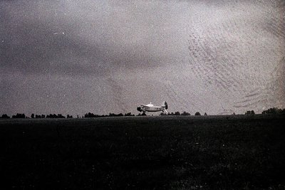 Vintage monochrome photo of a single-engine propeller aircraft taking off from a flat, open field. The plane’s high-wing desi...