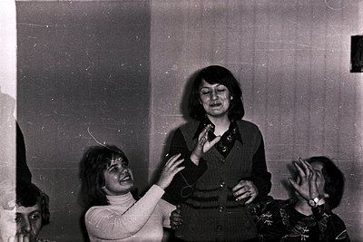Black-and-white candid shot of three women in a 1970s indoor setting, likely a social gathering. Central figure wears a patte...