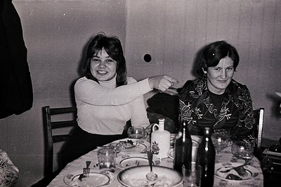 Two women seated at a set table in a dimly lit room, likely mid-20th century. The woman on left wears a turtleneck, smiling; ...