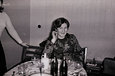 Vintage black-and-white photo of a seated woman in floral-patterned dress, holding a phone mid-conversation at a dining table...